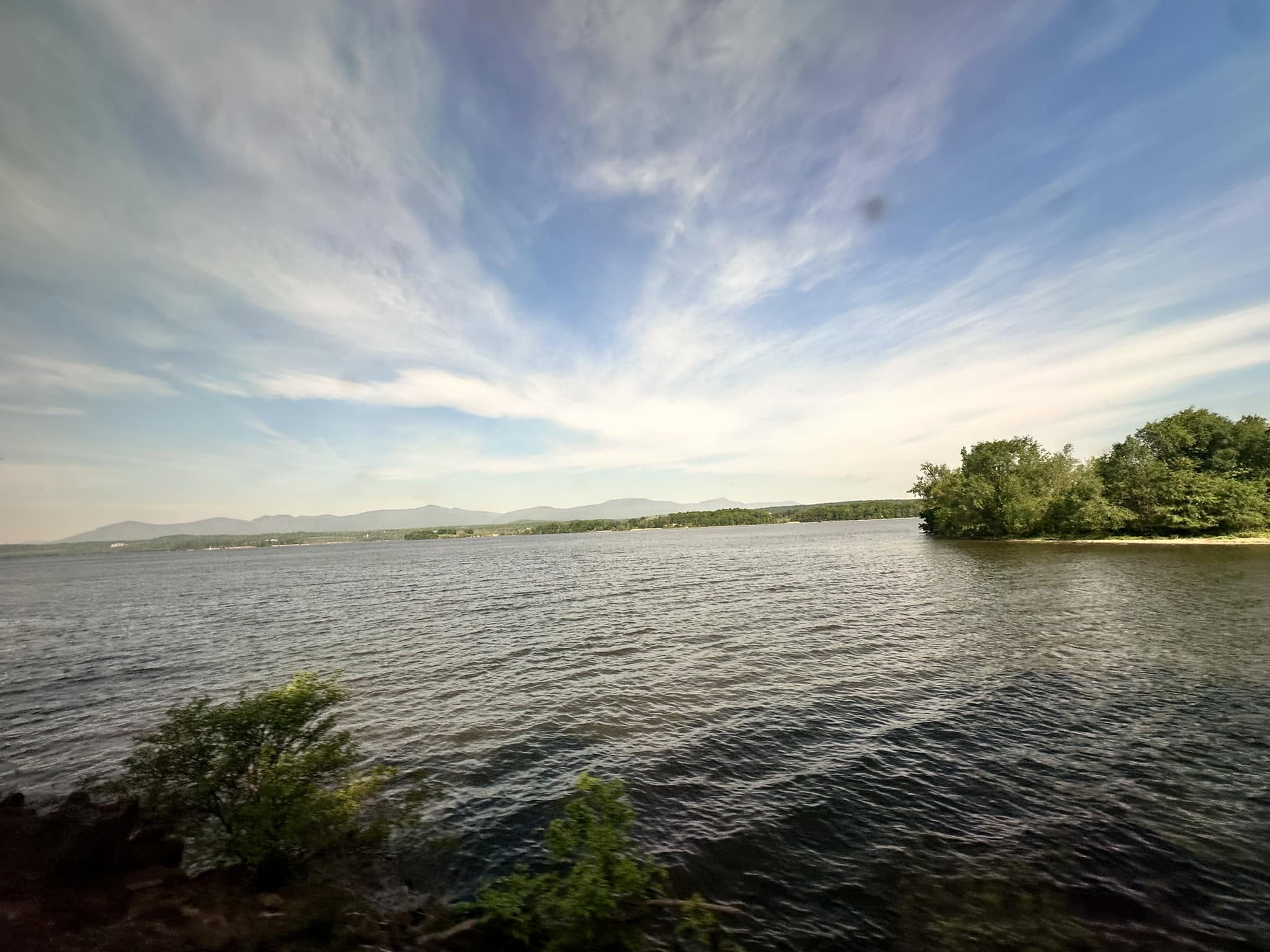 The Hudson River shown from the window of an Amtrak Cafe Car. The sky is clear with a few wispy clouds, 
            and the water has small waves. There are a few trees on an island and mountains in the distance, across 
            the river.